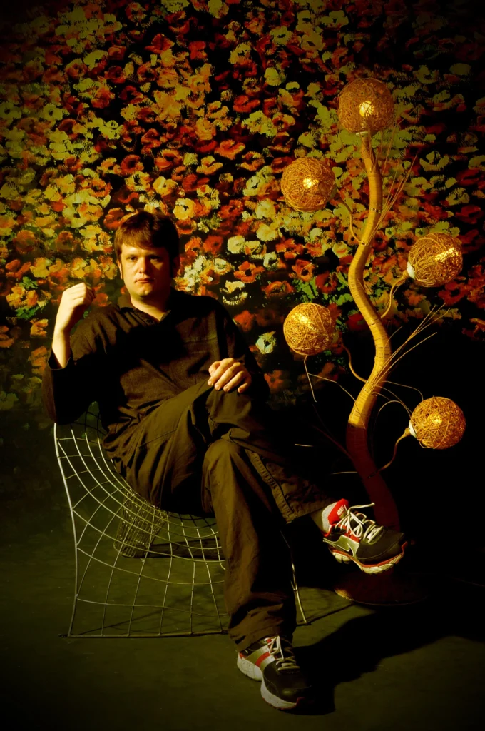 An old photo of Ozject in his mid-twenties, sitting in a wire chair with autumn colours and an abstract lamp beside him.