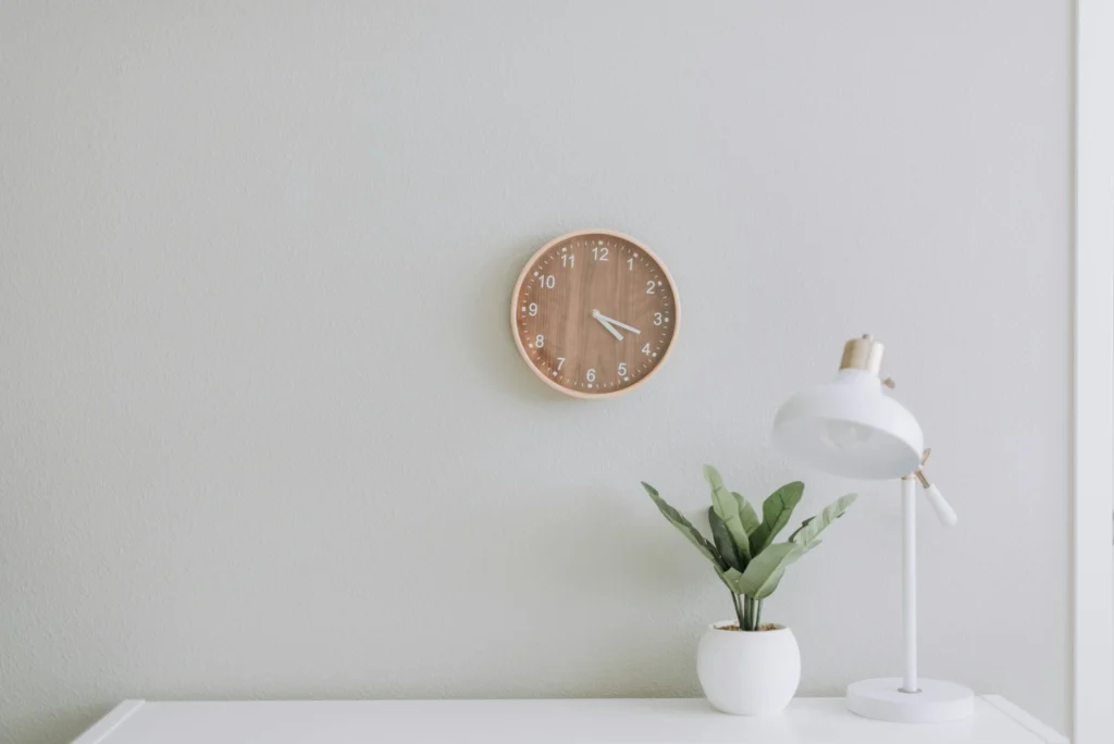 A stock photo of a minimalist desk with one plant, one lamp, and a clock behind the desk. A white desk with a white wall behind it.
